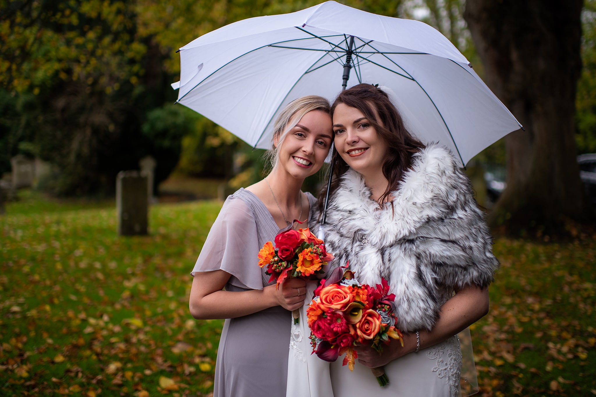 Bride & Bridesmaid sharing white umbrella, smiling and standing in church yard holding Autumn flower bouquets